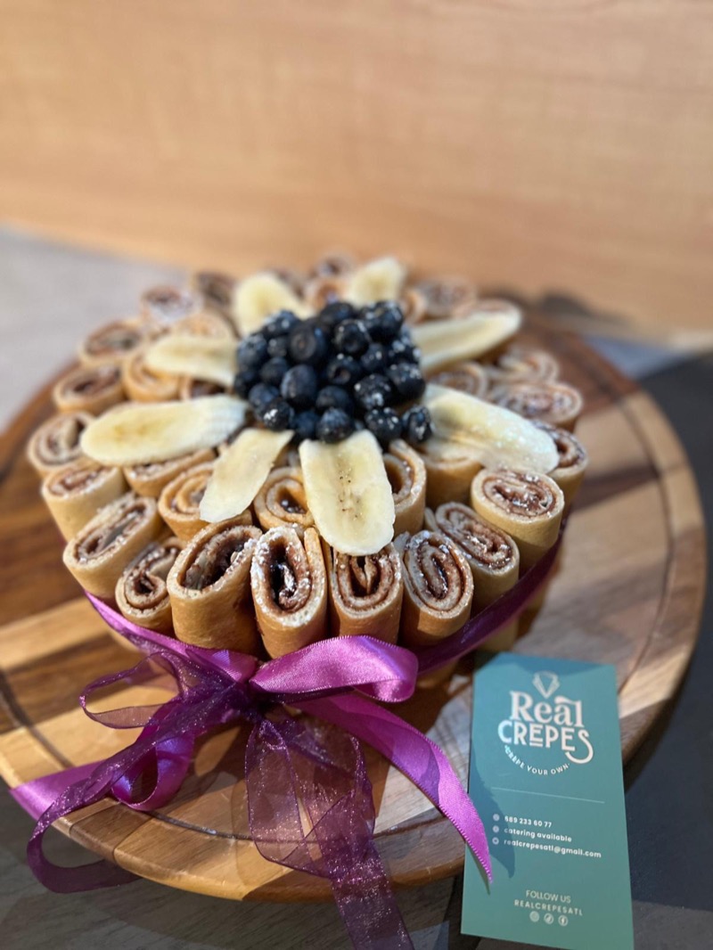 A Real Crepes crêpe cake of rolled sweet crêpes topped with banana slices and blueberries, tied with a ribbon.