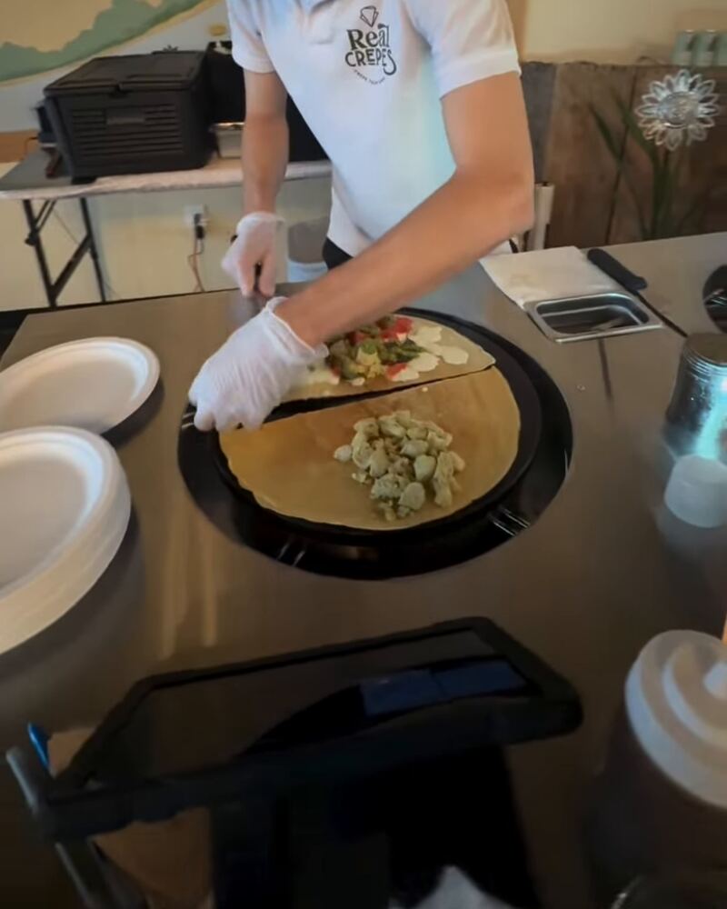 A savory crepe topped with chicken, tomato and herbs being folded on the griddle.
